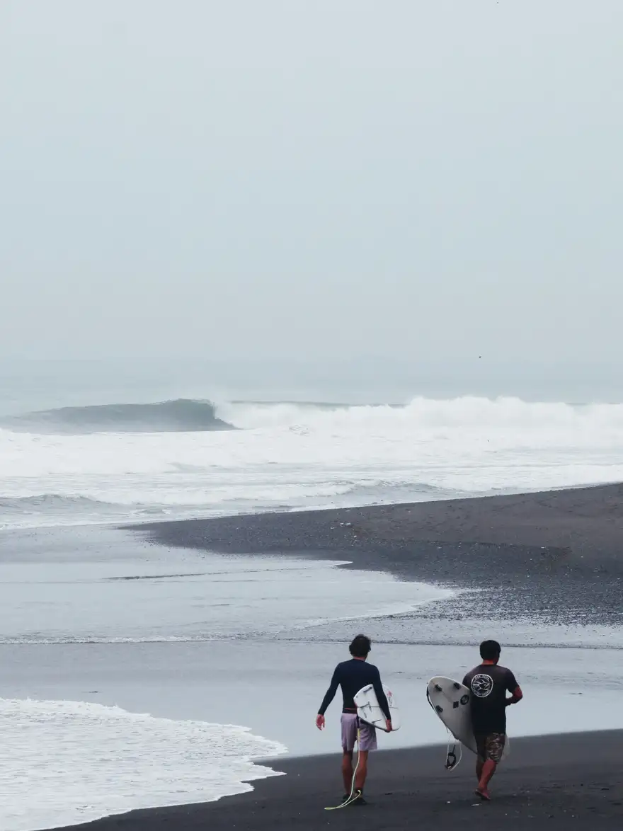 A surf coach and surfer walking down a black sand beach in Bali towards a peeling wave